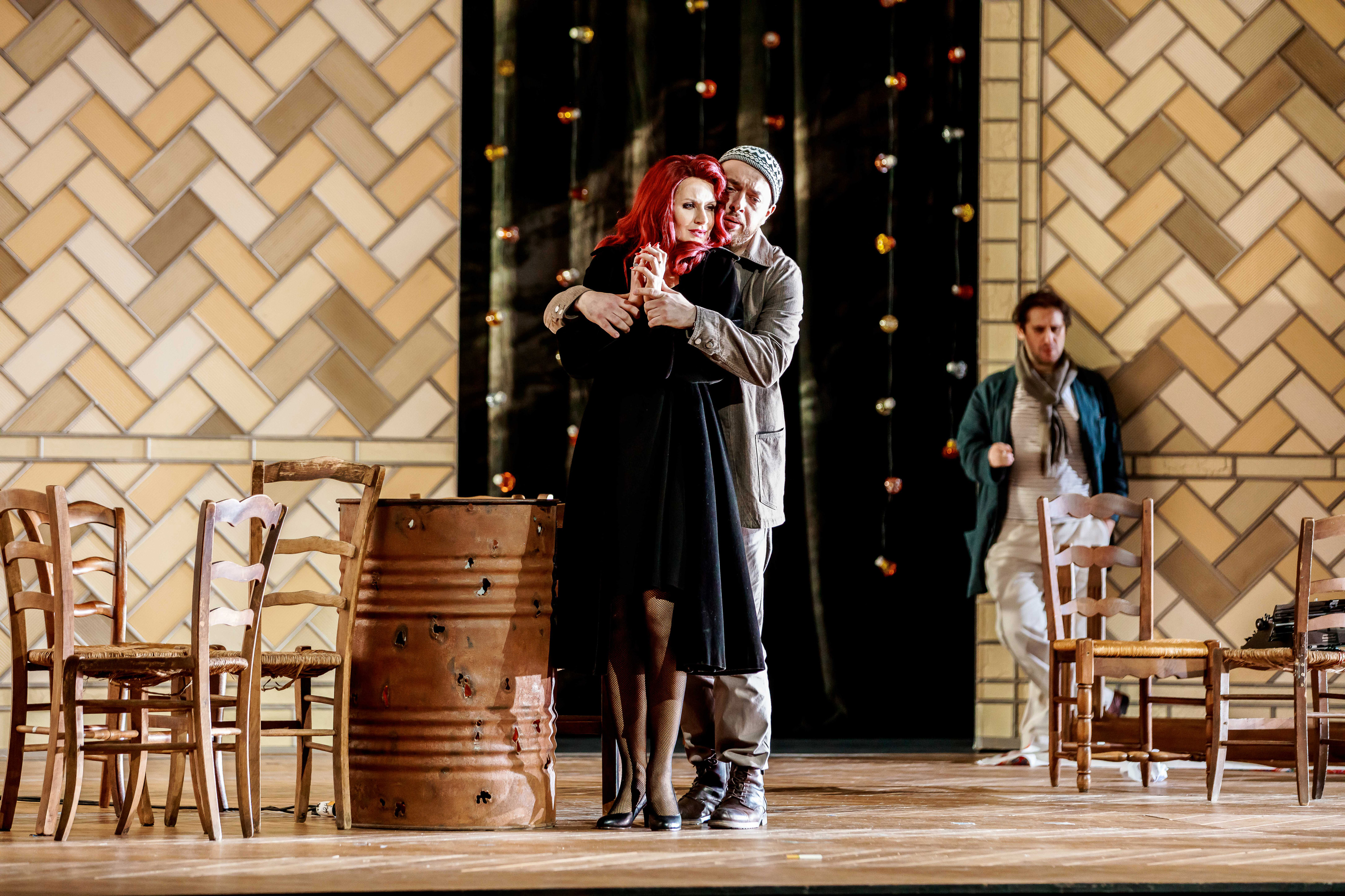 Edyta Piasecka (Fiorilla) in a black dress and long, red wig is hugged from behind by Łukasz Goliński (Selim) wearing a Turkish hat. Both are observed by Giulio Mastrototaro (Poet) leaning against a wall and holding a cigarette